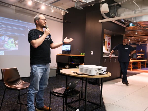 A man is giving a presentation or talk, holding a microphone and gesturing, while standing in front of a projected screen displaying text and digital artwork. The event takes place indoors in a modern venue with industrial ceilings and dark walls, and another man is visible standing in the background.