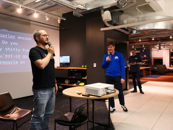 A photograph captures an indoor presentation or talk featuring three men, two of whom are holding microphones, standing in front of a projection screen displaying text in both French and English.