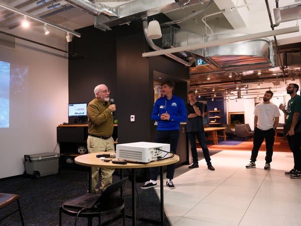 A candid photograph taken indoors shows an older bearded man giving a presentation using a microphone, standing next to a younger man in a blue jacket, while a projector screen displays an image of two dolphins underwater.