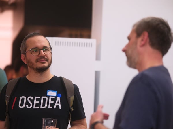 A candid photograph taken indoors showing a man wearing glasses and a black t-shirt with '/OSEDEA' printed on it, engaged in conversation with another man visible in profile on the right.