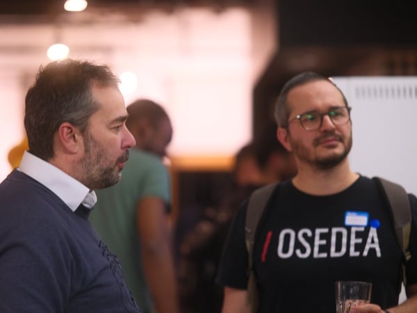 A candid photograph taken indoors at a professional or networking event, showing two men in the foreground, one of whom is wearing a black t-shirt with the text "/ OSEDEA".