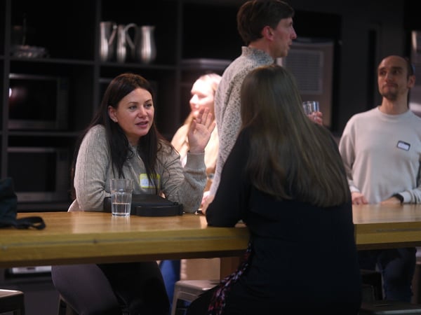 A candid photograph captures a group of five people socializing around a high wooden counter in a dimly lit, modern indoor space, with one woman in the foreground actively talking and gesturing with her hand.