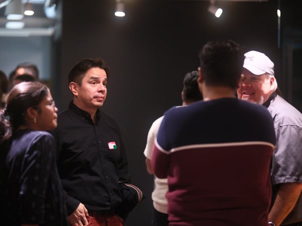 A candid photograph captures a group of people socializing and conversing indoors in a dimly lit venue, focusing on a man in a black shirt and red pants standing in profile next to a woman.