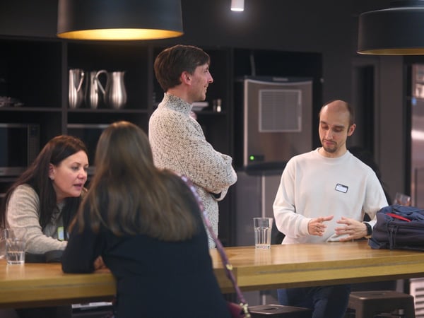 A candid photograph captures four people gathered around a wooden counter in a modern, dark-toned office break room or kitchen area, engaged in a casual discussion.