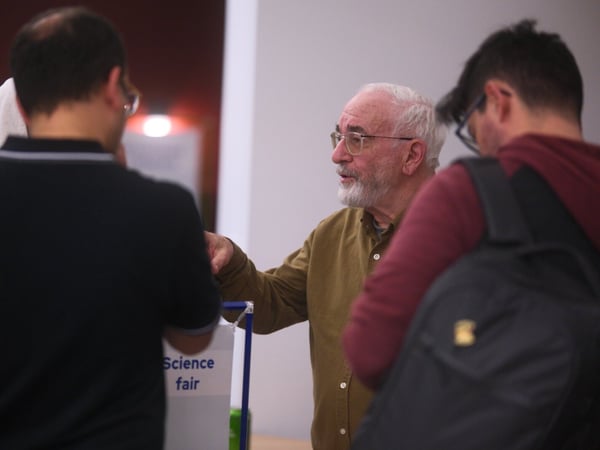 An older man with a white beard and glasses, wearing a khaki shirt, stands engaged in conversation with several younger attendees at an indoor event, likely a science fair, as suggested by a partially visible sign.