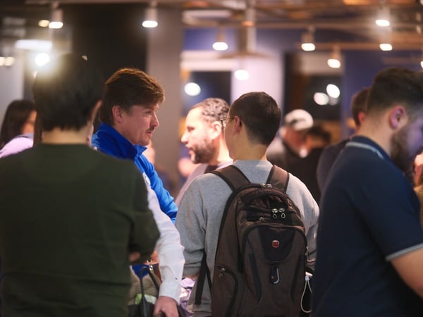A candid indoor photograph captures a group of people mingling and engaged in conversation in a dimly lit venue, focusing on a man in a blue jacket talking to two individuals whose backs are mostly toward the camera.