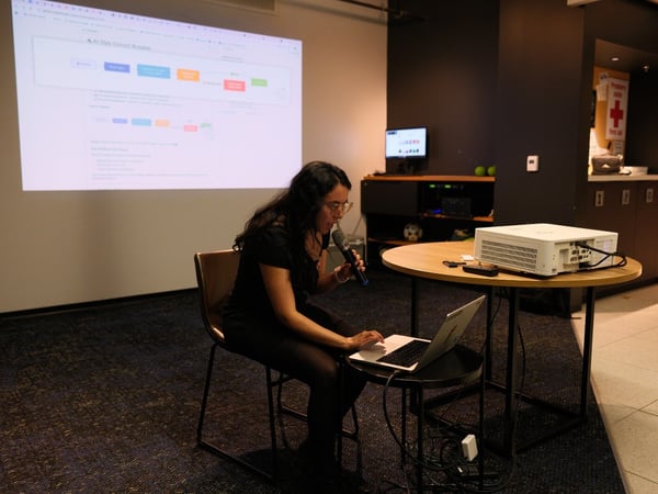 A woman is seated indoors, giving a technical presentation or demo, speaking into a microphone while operating a laptop, with a diagram titled 'AI Ops Circuit Breaker' projected onto a large screen behind her.