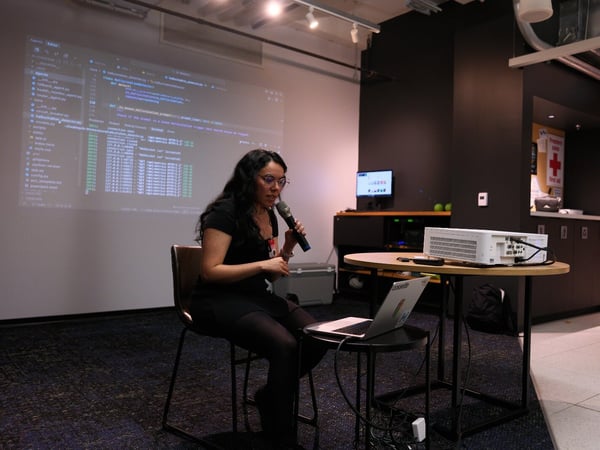 A woman is seated on a chair holding a microphone, giving a technical presentation in a dimly lit indoor venue, with a large screen displaying lines of programming code behind her.