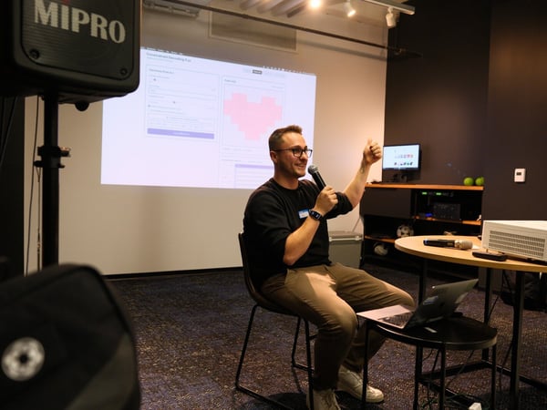 A man wearing glasses is giving a presentation, sitting down while holding a microphone and gesturing a thumbs up in front of a projection screen displaying a web interface featuring forms and a large pixelated red heart.