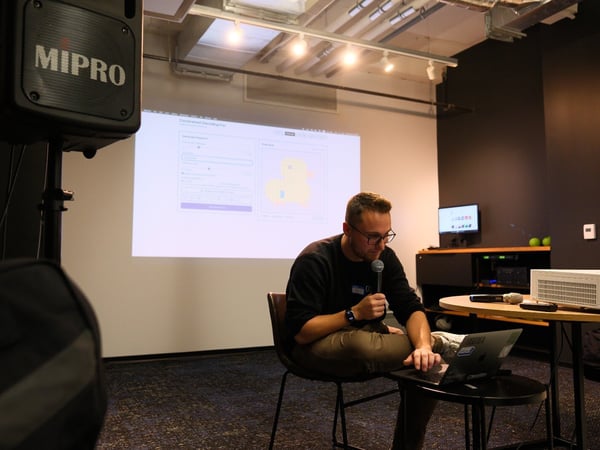 A man wearing glasses is seated, holding a microphone and interacting with a laptop during an indoor presentation or talk. Behind him, a large screen displays a web application interface next to a prominent MIPRO speaker on the left.