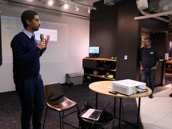 A photograph capturing an indoor presentation where a man, dressed in a navy sweater and collared shirt, stands holding a microphone and gesturing in front of a projected screen showing data; another man stands observing near a dark wall on the right.