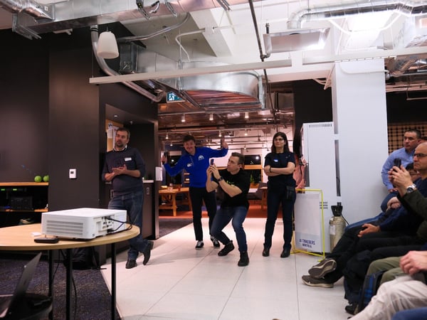A candid photograph captures an indoor event or presentation in a modern office space, showing a small group of people standing near a projector and table while others are seated in the audience.
