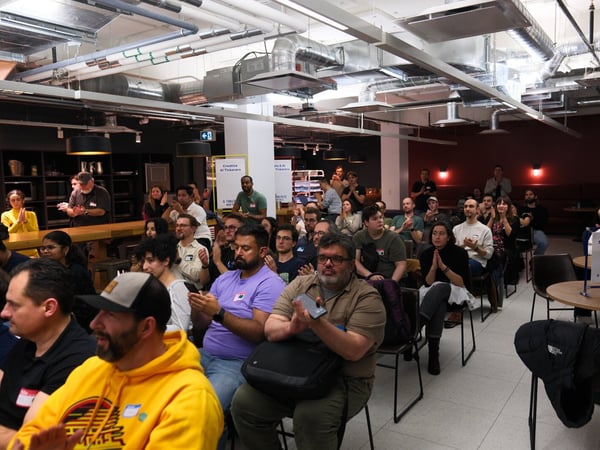 A candid photograph captures a large, diverse audience seated indoors at an event, with most attendees clapping in applause, suggesting the conclusion of a presentation or talk.
