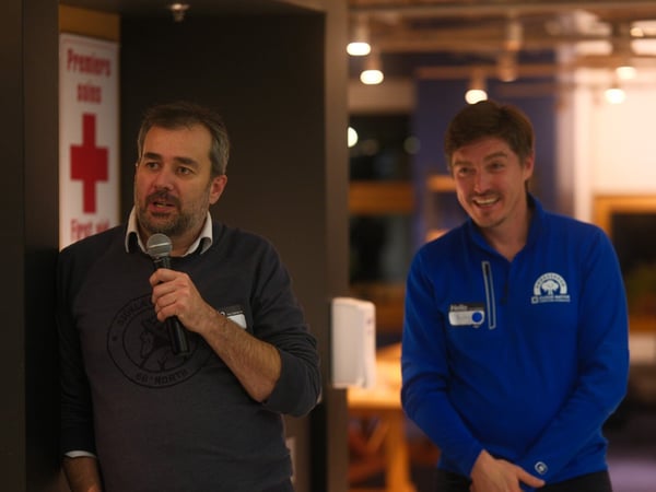 A candid photograph captures two men standing indoors at an event, where one man on the left is speaking into a microphone while the man on the right smiles and looks on. A bilingual sign for first aid ('Premiers soins' and 'First aid') is visible behind the speaker.