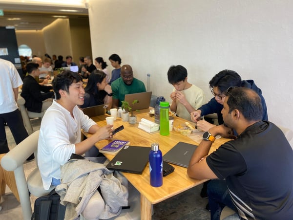 A group of people are gathered around a large wooden table in a modern indoor workspace or cafe, engaged in various activities like working on laptops, eating, and conversing.