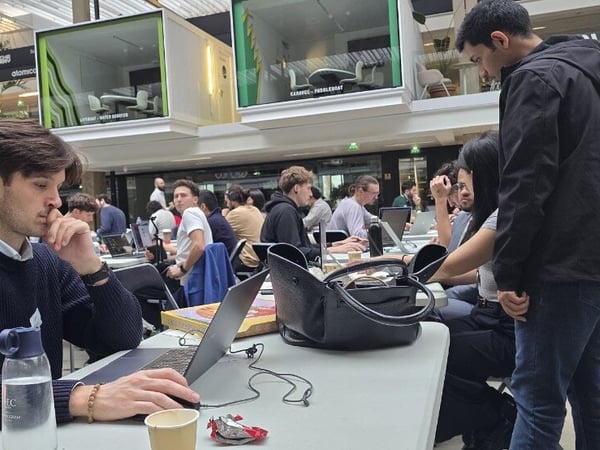 A candid photograph captures a busy indoor tech event or co-working session, showing many attendees working intently on laptops at long tables in a modern atrium setting, with prominent "Google for Startups" signage visible in the background.