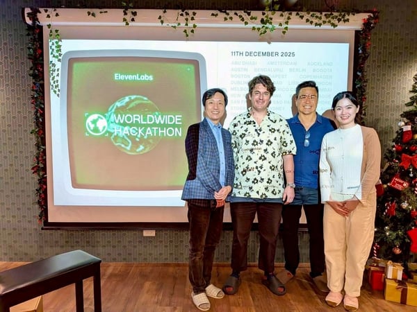 Four individuals, three men and one woman, pose smilingly in front of a projection screen advertising the 'ElevenLabs Worldwide Hackathon' scheduled for December 11, 2025, in a room decorated with Christmas garlands and a Christmas tree.