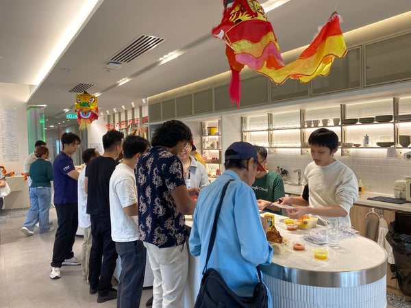 A group of people are gathered around a counter in an office kitchen decorated with traditional Chinese lion dance heads, likely for a social or festive event.