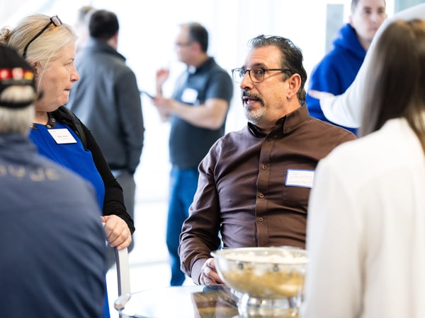 A candid photograph of people socializing at an indoor event, with a man in a brown shirt and glasses engaged in conversation in the foreground.