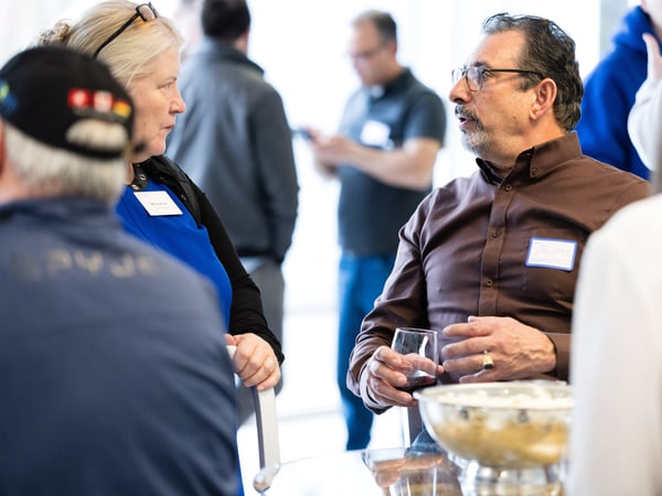 A group of people engaged in conversation at a social or networking event, with a man in a brown shirt speaking while holding a glass of wine.