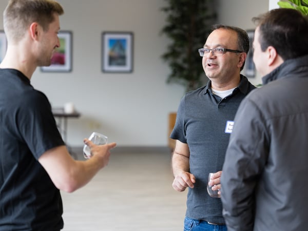 Three men are standing in a large indoor space, engaged in a social conversation while holding glasses. One man faces the camera directly, while the others are seen from the side or back.