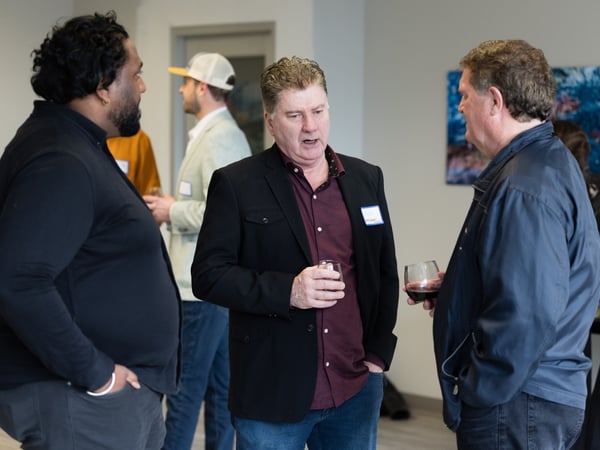 A group of people are gathered at an indoor social event, with three men in the foreground engaged in conversation while holding drinks.
