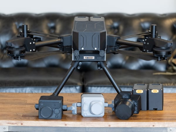 A large industrial drone sits on a wooden table alongside several modular camera payloads and spare batteries, with a leather sofa in the background.
