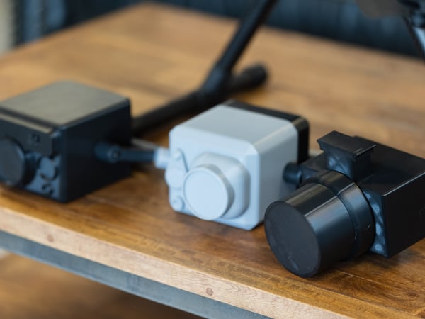 A close-up photograph of three small, cube-shaped electronic devices, likely sensors or cameras, resting on a wooden table.