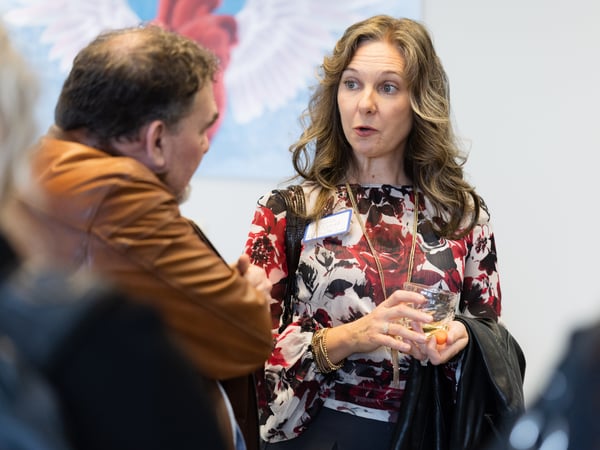 A woman in a floral blouse holding a drink is engaged in a conversation with a man at a social gathering.