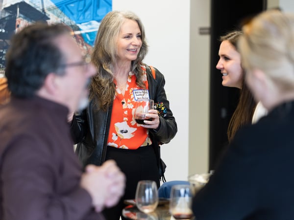 A group of people are socializing at an indoor event, with a woman in a red floral shirt and black leather jacket smiling and holding a glass of wine in the center.