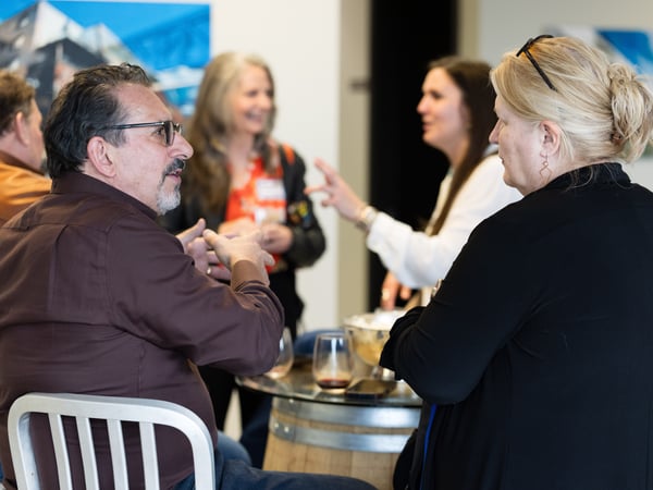 A group of people are engaged in a lively conversation at an indoor social gathering, likely at a winery or tasting room.