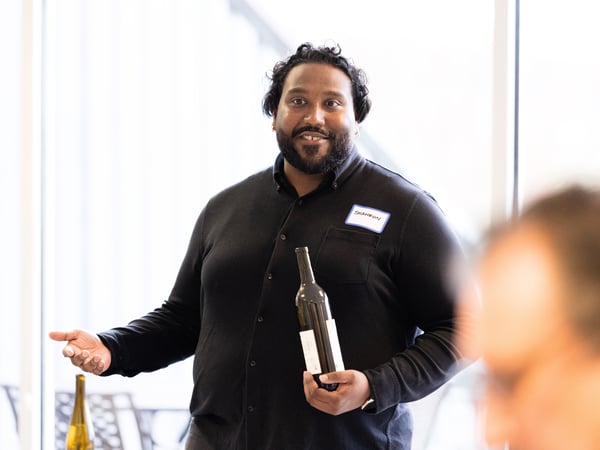 A man in a black button-down shirt holds a wine bottle while gesturing, appearing to be giving a presentation or talk in a bright indoor setting.