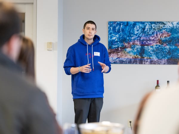 A man in a blue hoodie is giving a presentation or speaking to a group of people in an indoor setting.