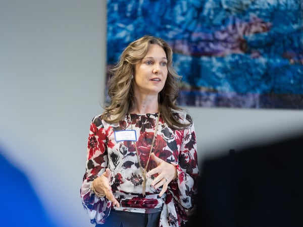 A woman with long wavy hair, wearing a floral print blouse, is speaking and gesturing with her hands in an indoor setting.