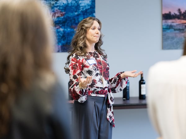 A woman with long wavy hair, wearing a red and white floral blouse, stands and gestures while speaking to a group of people in an indoor setting.