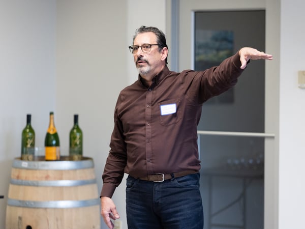 A man in a brown shirt and glasses is captured in an indoor setting, gesturing with his left arm. In the background, several wine bottles are placed on top of a wooden barrel.