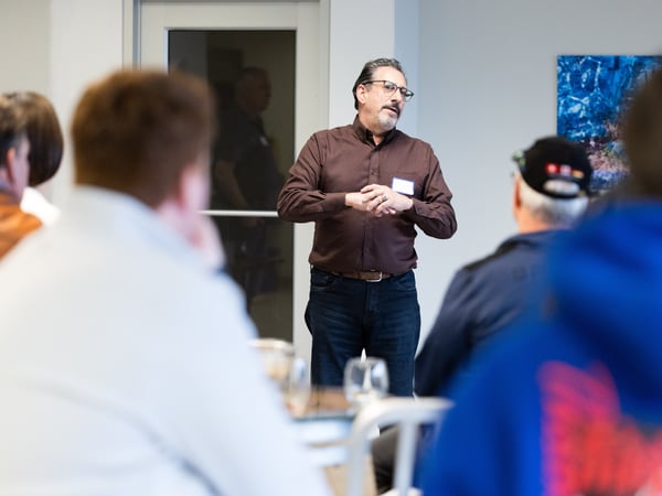 A man in a brown shirt and glasses is speaking to a group of people in an indoor setting, appearing to lead a presentation or discussion.