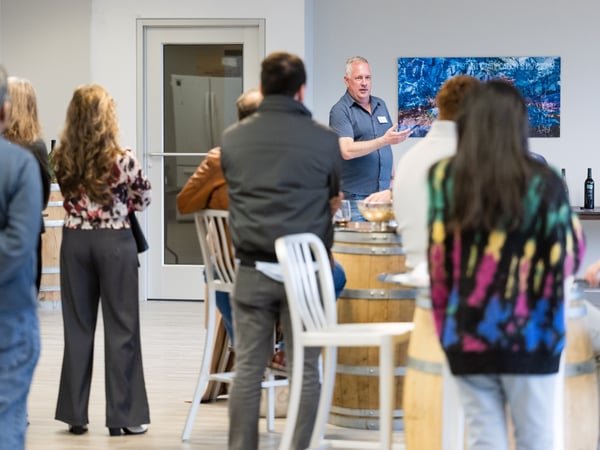 A man stands in an indoor space, possibly a winery, giving a presentation or speech to a group of people who are mostly seen from behind.