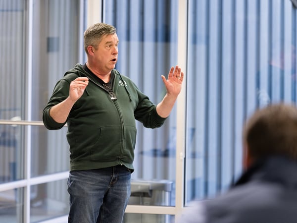 A man in a green zip-up hoodie and blue jeans stands in an indoor space with large glass windows, gesturing with both hands as if speaking or giving a presentation.