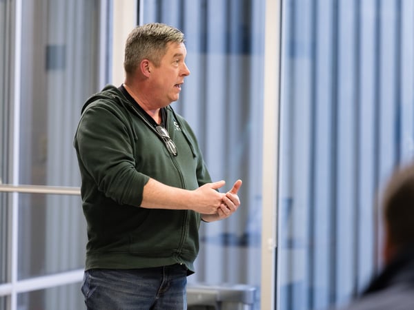 A man in a green zip-up hoodie and jeans is captured in profile, speaking and gesturing with his hands in an indoor setting with large windows.