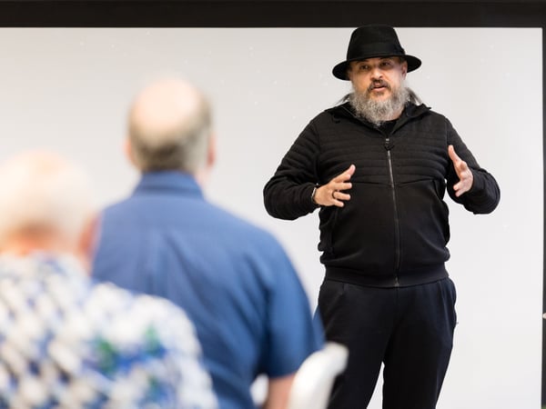 A man with a beard and a black hat is gesturing with his hands while speaking to an audience, with two blurred figures in the foreground.