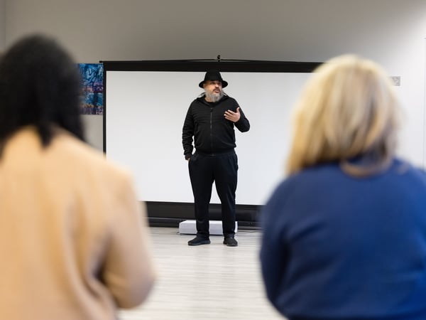 A man in a black hat and outfit stands in front of a projector screen, giving a presentation to an audience.