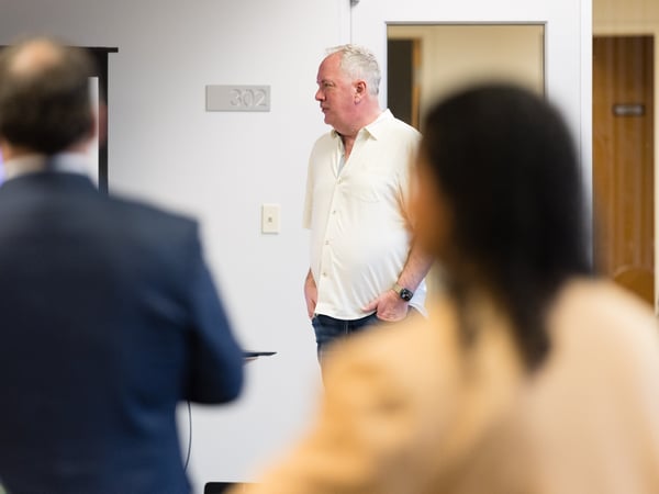 A man in a light-colored button-down shirt stands in an indoor setting, likely a classroom or office, looking towards a presentation screen while other people are blurred in the foreground.