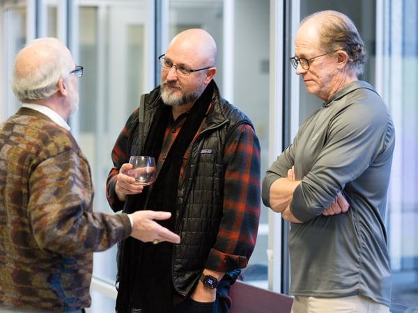 Three older men are standing in a modern indoor space with glass partitions, engaged in a conversation.