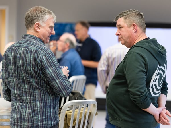 A candid photo of two men engaged in conversation at an indoor social event, with other guests visible in the blurred background.