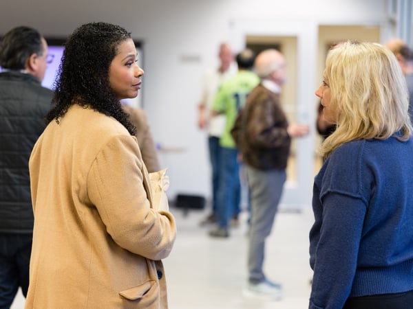 A woman with curly dark hair in a tan coat is engaged in a conversation with a blonde woman in a blue sweater in an indoor setting.