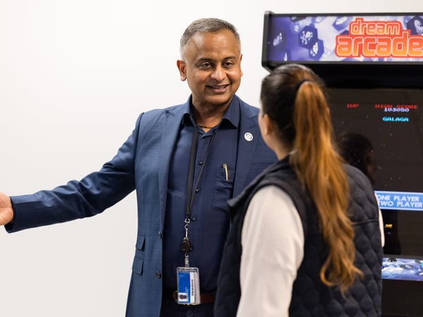 A man in a blue suit is gesturing while talking to a woman standing in front of a retro 'dream arcade' machine.