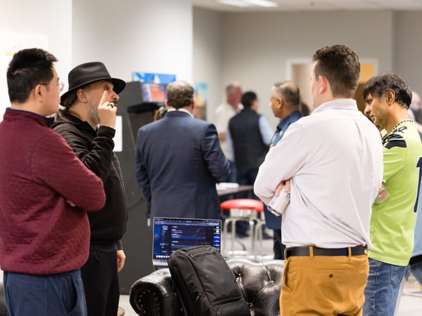 A group of men are gathered in an indoor office or event space, engaged in a conversation. One man wearing a black hat is gesturing with his finger while talking to others.