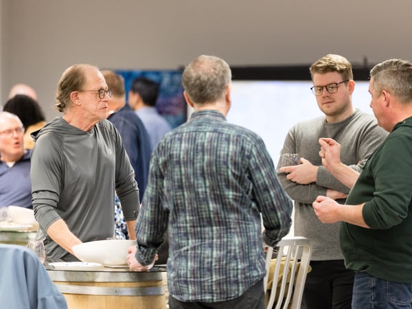 A group of people are engaged in conversation at an indoor social or networking event, with some standing around a barrel used as a table.