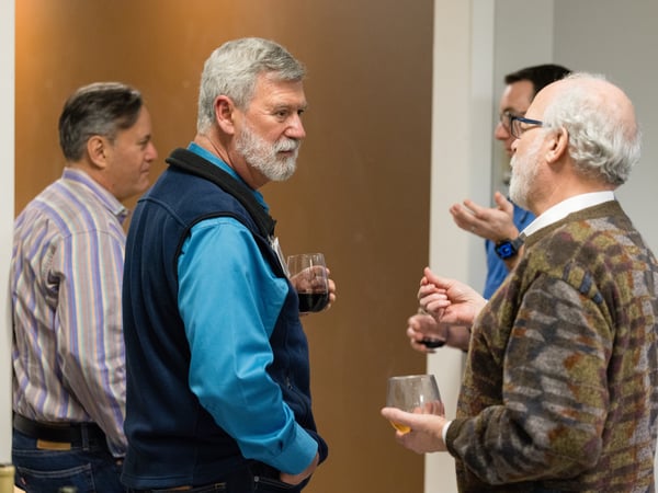 A group of men are engaged in conversation during an indoor social gathering, with some holding glasses of wine.